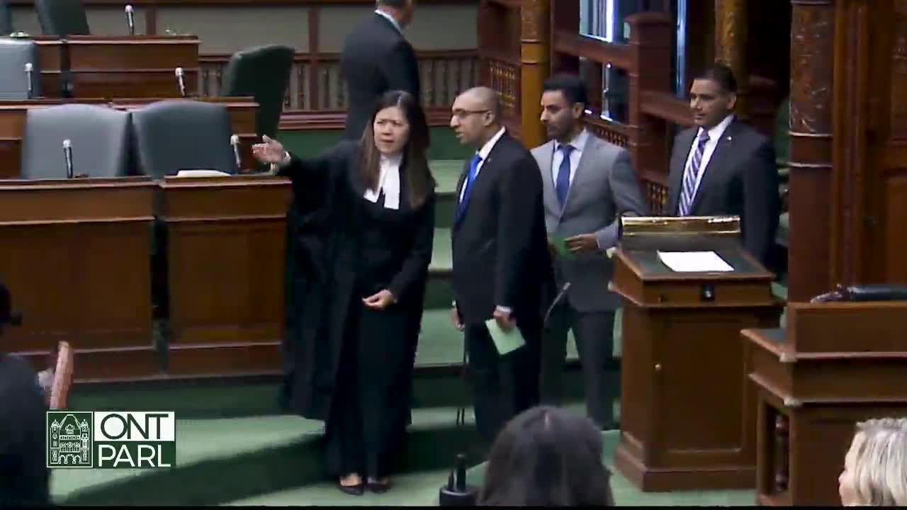 In the Legislative Assembly of Ontario, a woman in a black robe gestures with her arm, directing the attention of the men beside her. One of the men, holding a green paper, looks on as they move toward a podium.
