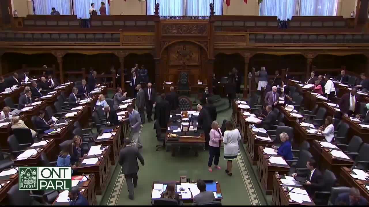 People are milling about the floor of the Legislative Assembly of Ontario, some in suits, others in more casual attire. A few individuals are gesturing animatedly, while others are heading toward the front.
