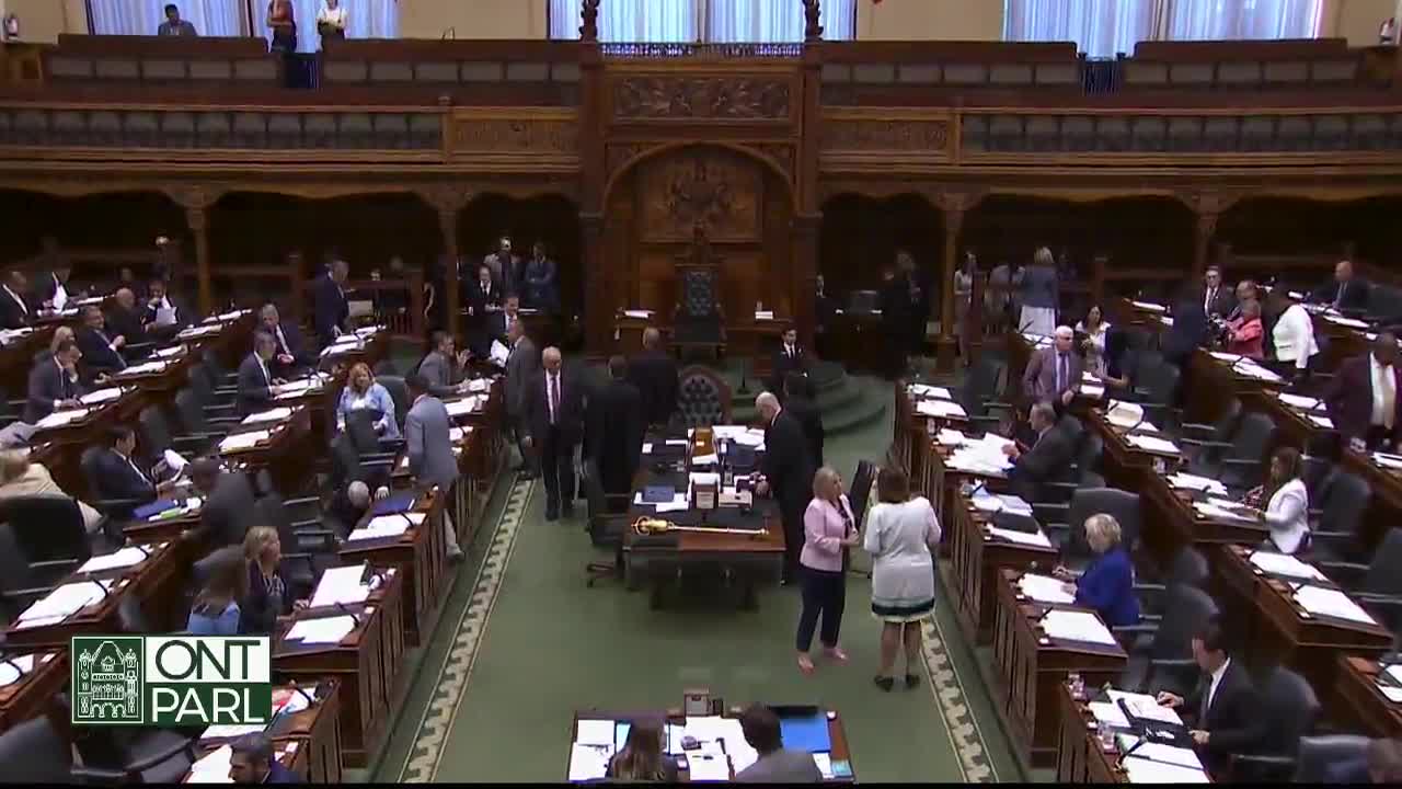 People in suits and dresses mill about the floor of the Legislative Assembly of Ontario, some chatting, others heading toward their seats. A few figures gather near the Speaker's table, the iconic gavel resting on the desk.
