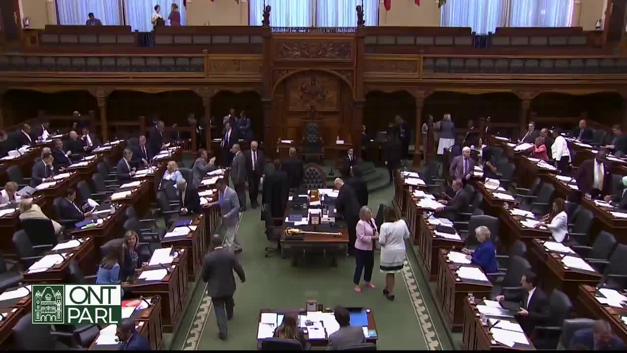 People in suits and dresses mill about the floor of the Legislative Assembly of Ontario, some chatting and others heading toward the front. A few individuals are seated at their desks, seemingly preparing for a session.
