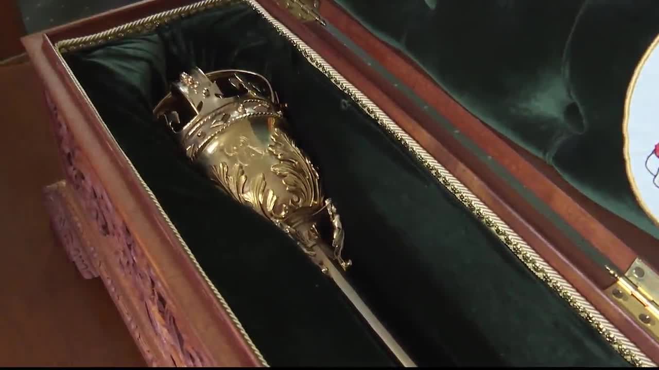 A golden mace, the symbol of the Legislative Assembly of Ontario, rests inside a velvet-lined case. The ornate details of the mace's crown and shaft gleam against the dark green fabric.
