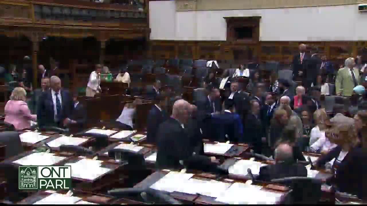 The Legislative Assembly of Ontario is abuzz with activity; people in suits and dresses mill about the chamber. Some are standing near the desks, while others are walking towards the back.
