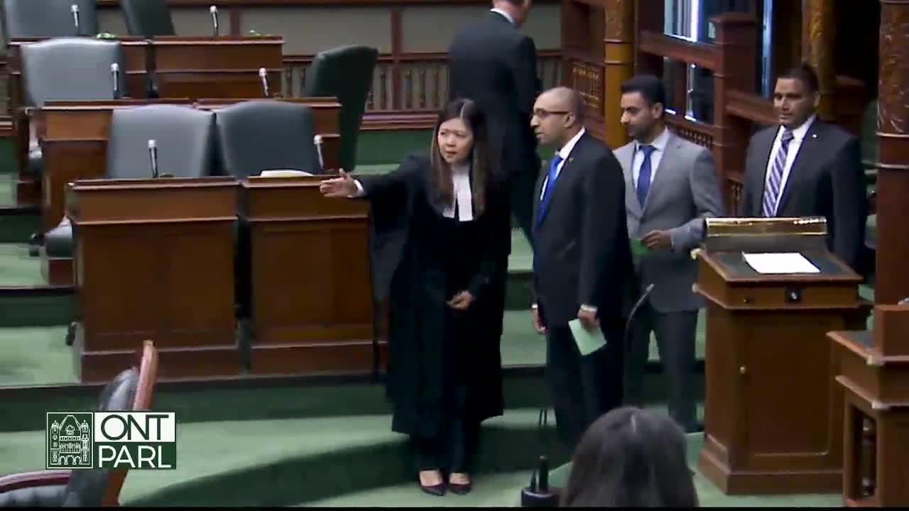 A woman in a black robe gestures with her arm extended, likely addressing the Legislative Assembly of Ontario. Several men in suits stand nearby, some holding papers.
