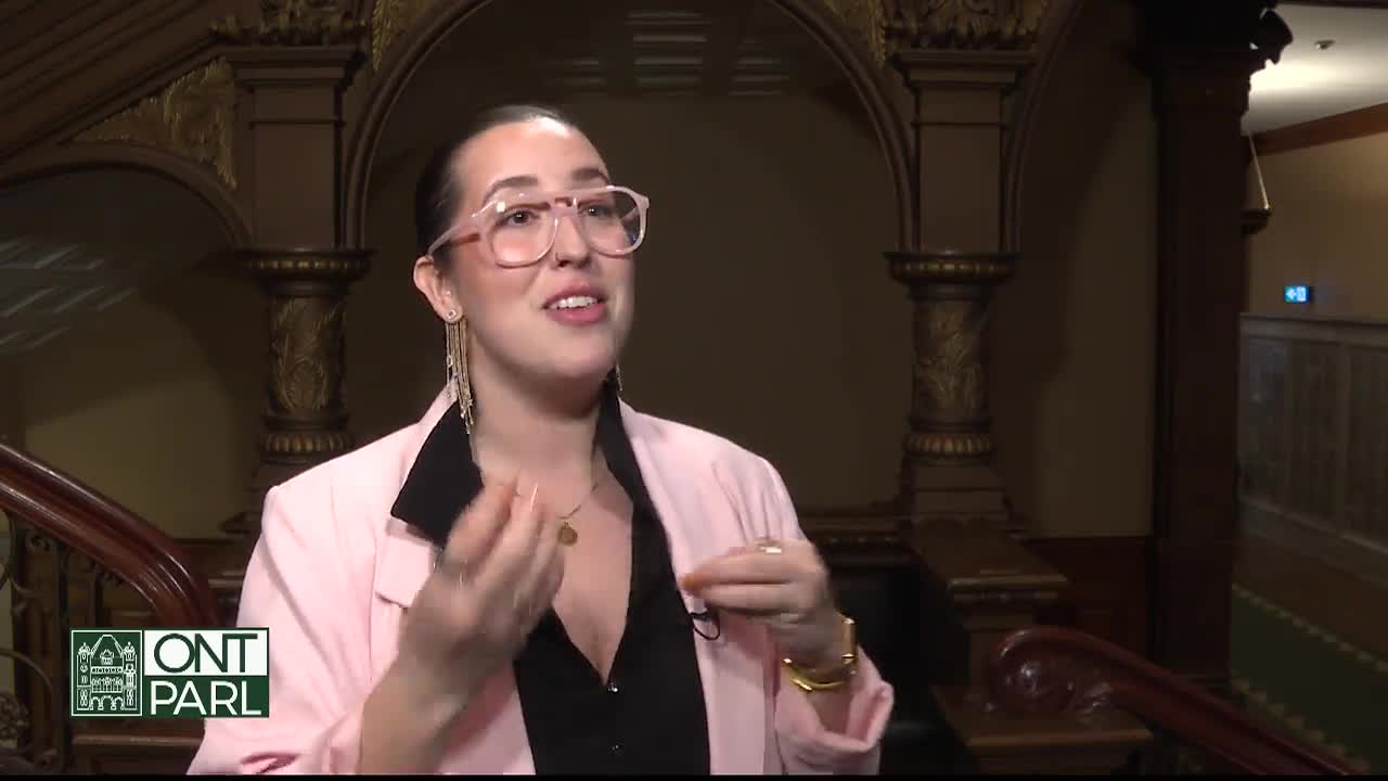 A woman in a pink blazer gestures with her hands as she speaks, likely in the Ontario Legislative Assembly. The ornate staircase and logo in the corner suggest this is a broadcast from the building.
