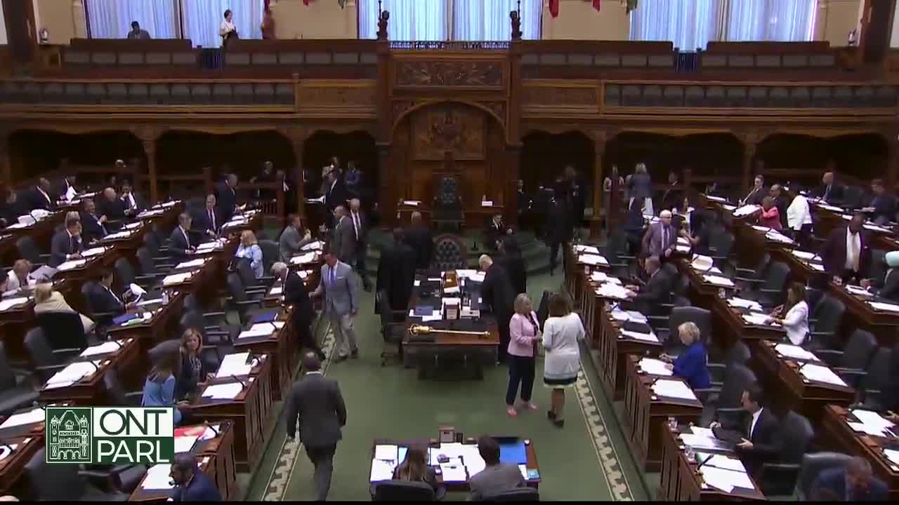 People in suits and dresses mill about the Legislative Assembly of Ontario chamber, some chatting near their desks. Several individuals walk in the aisles, heading toward the center of the room.

