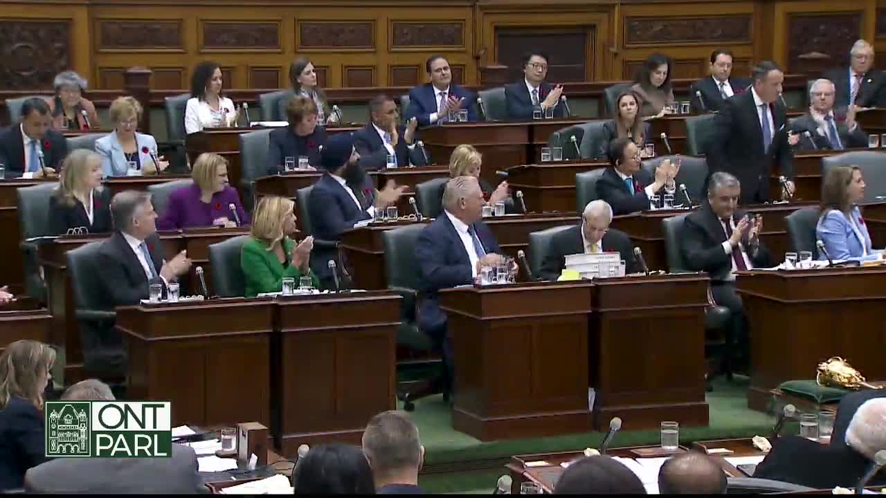 In the Legislative Assembly of Ontario, many members are clapping, while one man in a suit walks toward the front. Premier Ford sits at his desk, watching.

