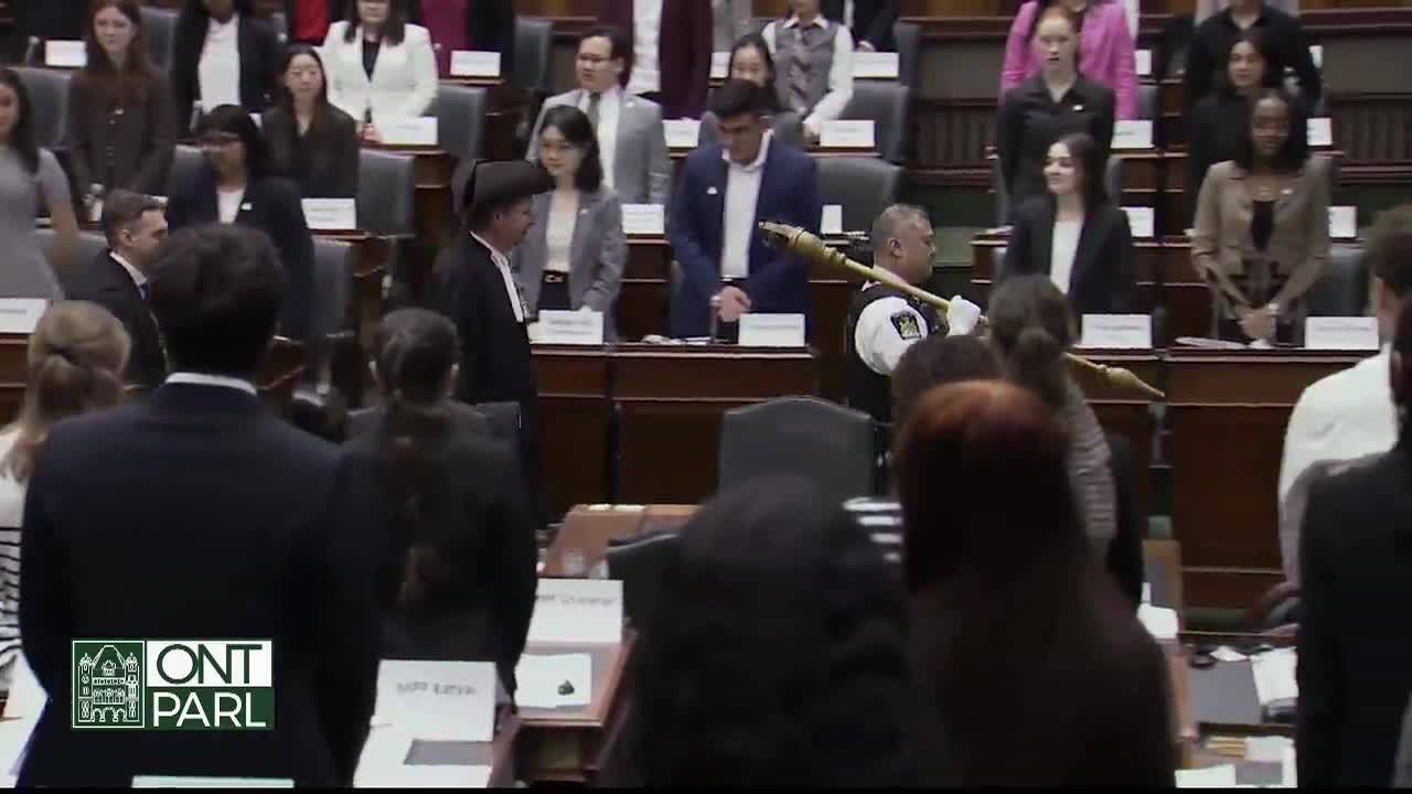 In the Legislative Assembly of Ontario, a man in a uniform carries a ceremonial mace through the room. The Speaker, in traditional robes, is also present as members of the assembly stand at their desks.

