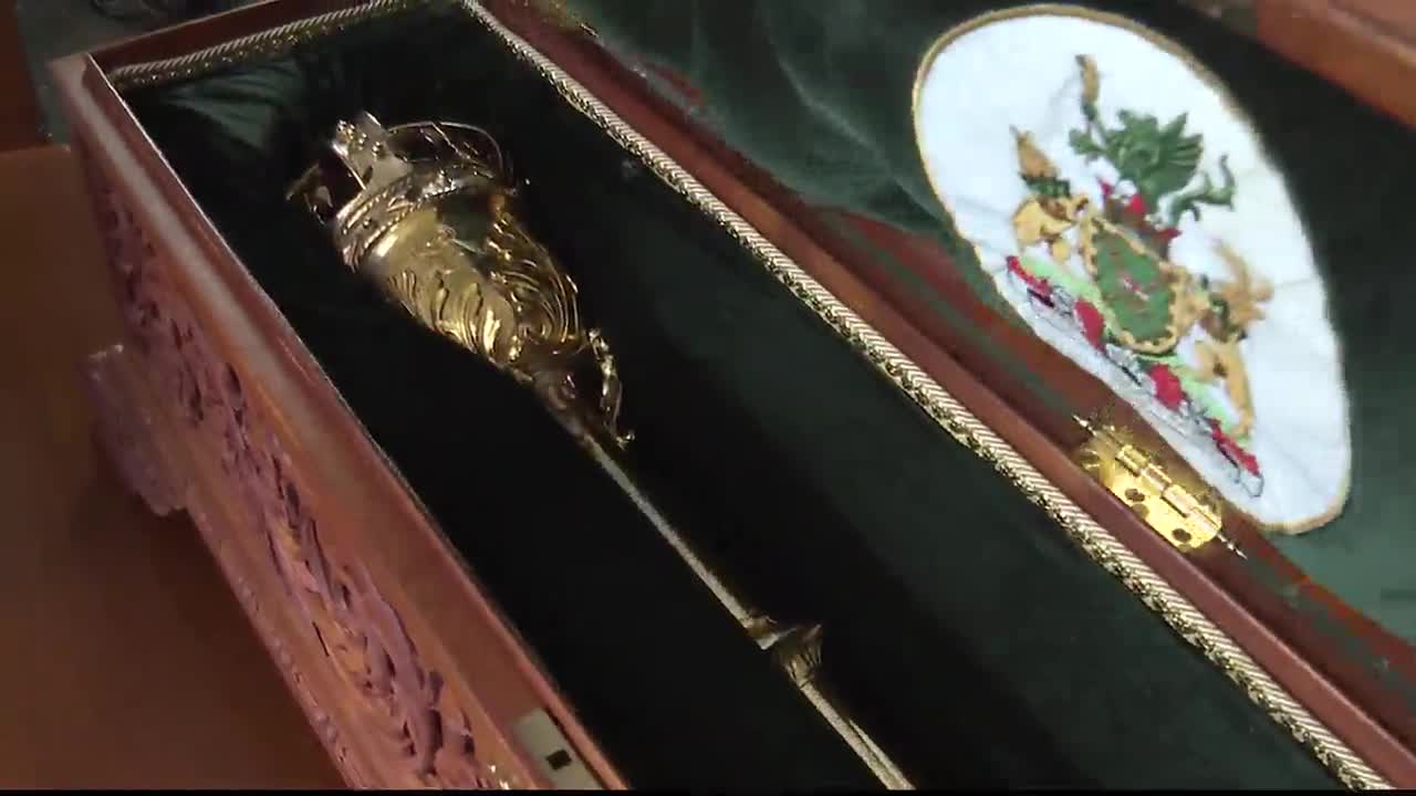 Inside a wooden case, the golden mace of the Legislative Assembly of Ontario rests on a bed of green velvet. Beside it, a small embroidered crest is visible.
