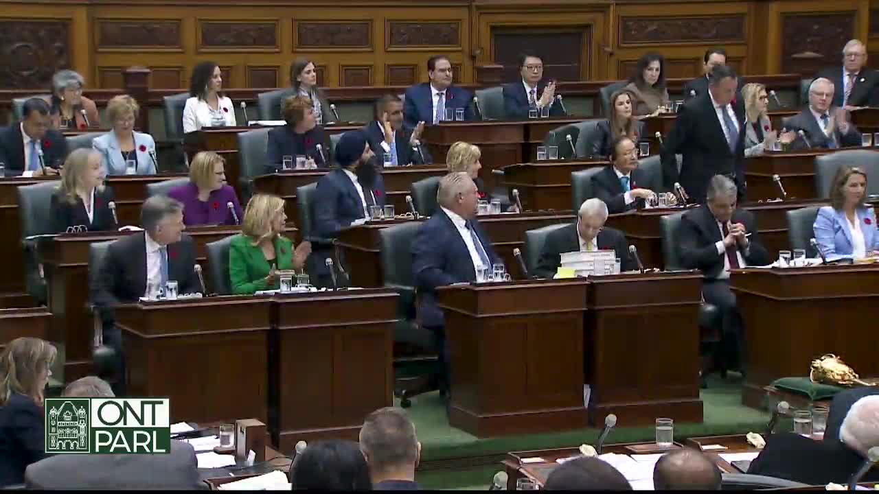 In the Ontario Legislative Assembly, a suited man strides down the aisle, while others clap. Premier Ford sits at his desk, watching the scene unfold.
