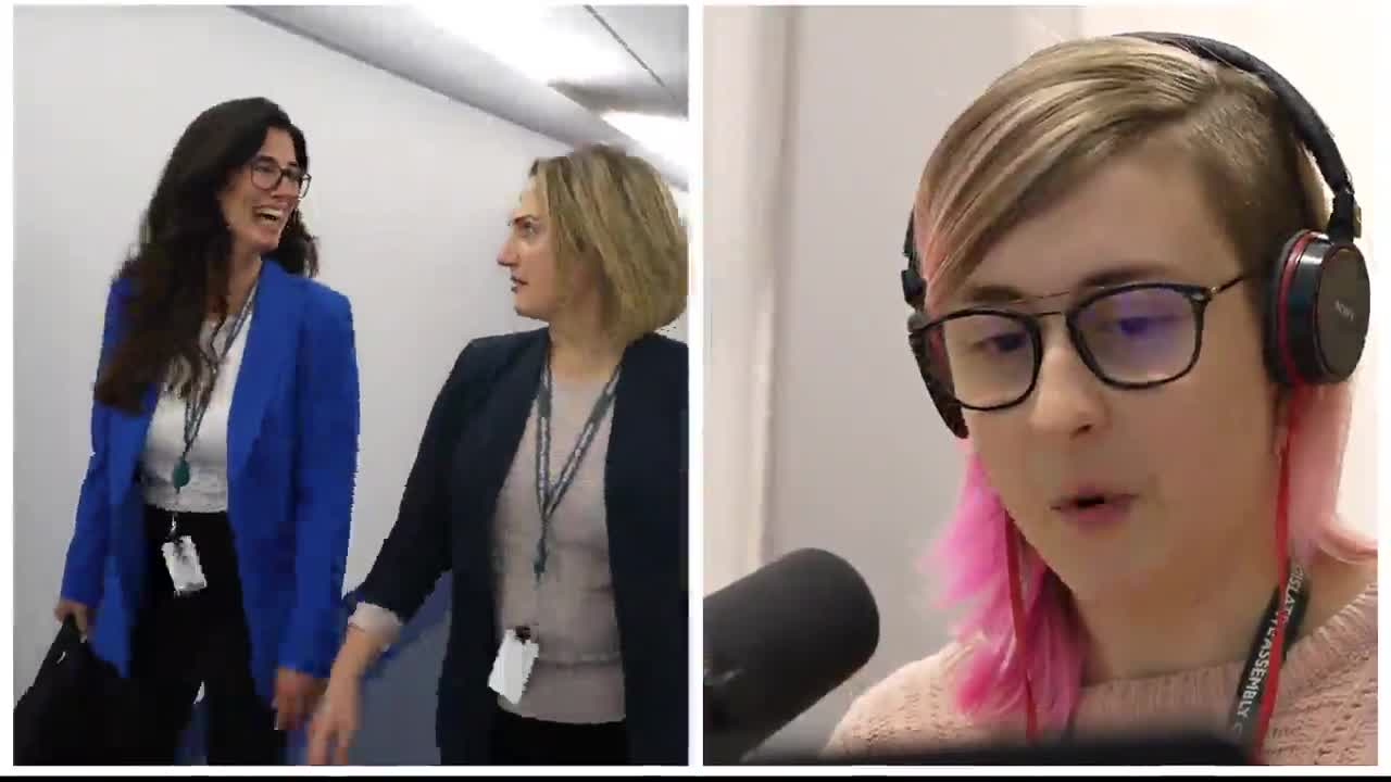 Two women in business attire are walking down a hallway, one laughing and the other looking ahead. On the right, someone with pink-streaked hair is speaking into a microphone, wearing headphones with the Legislative Assembly of Ontario lanyard.
