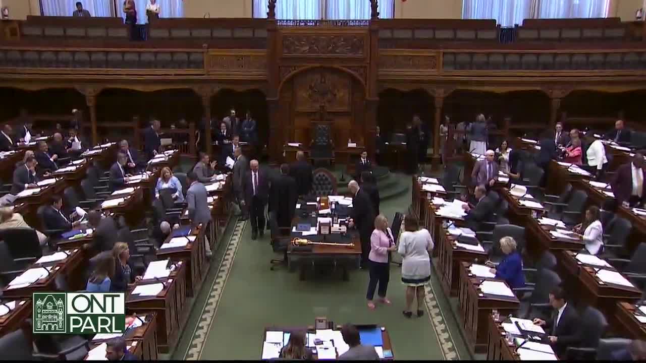 People in suits and dresses mill around the chamber, some chatting while others gather near the central table. The Ontario Legislative Assembly is in session, and the scene feels busy.
