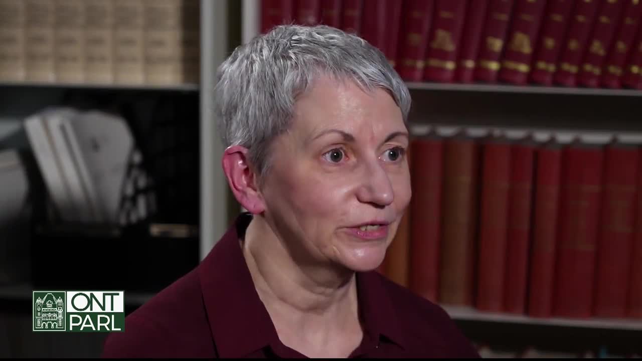 A woman with short, gray hair speaks directly to the camera, her expression earnest. Behind her, a shelf is lined with what appear to be red-bound books, likely within the Legislative Assembly of Ontario.
