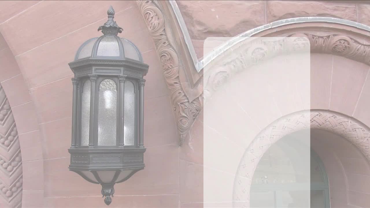 A detailed, ornate lamp hangs on the exterior of the Ontario Legislative Assembly. The building's pink stone facade is decorated with carved details and arched windows.
