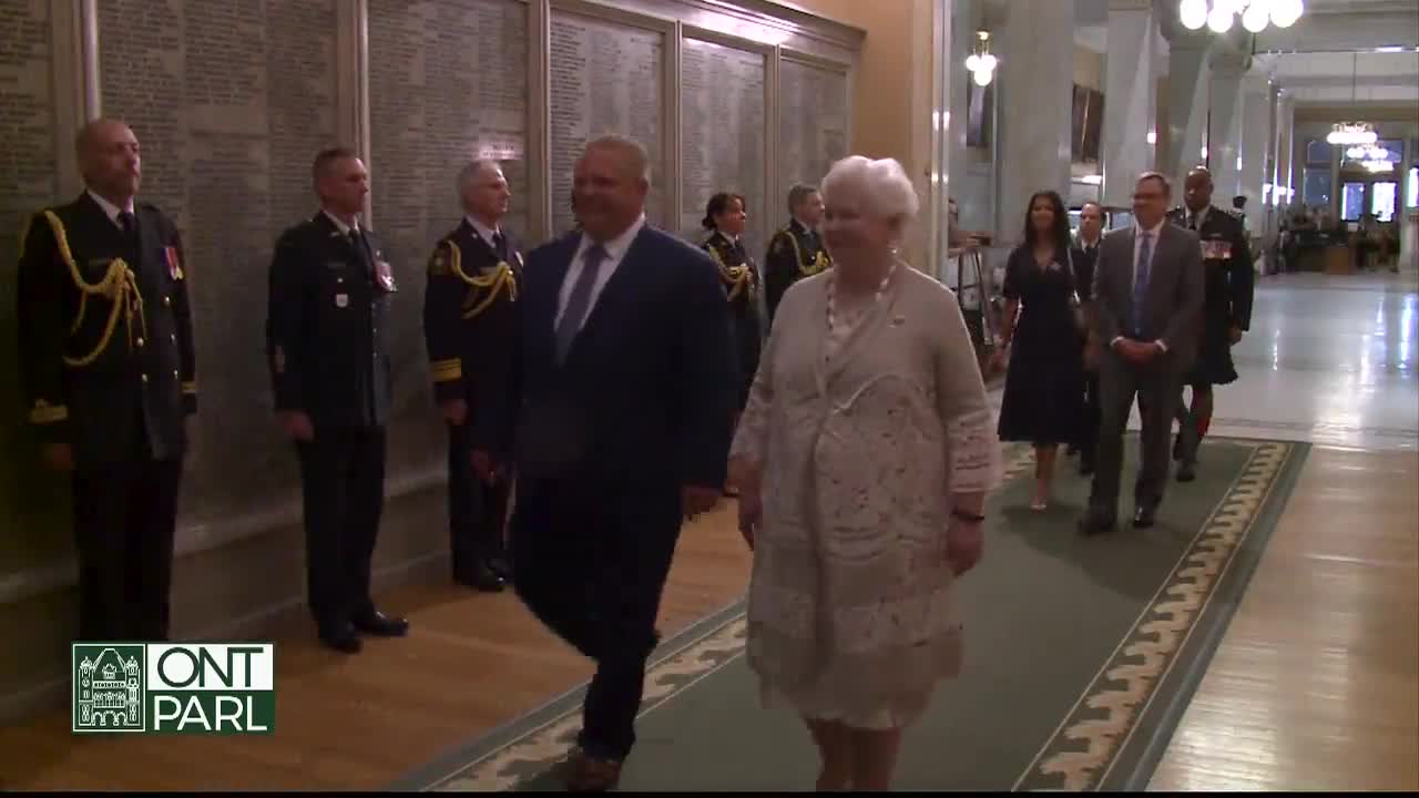 A man in a dark suit and a woman in a cream-colored dress walk side-by-side down a long hallway. Behind them, a small procession follows, including uniformed officers and other officials, all moving forward.
