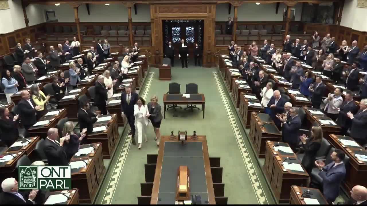 A woman in a white suit, flanked by others, walks down the central aisle of the Legislative Assembly of Ontario. The members of the assembly, in suits and dresses, are standing and applauding.
