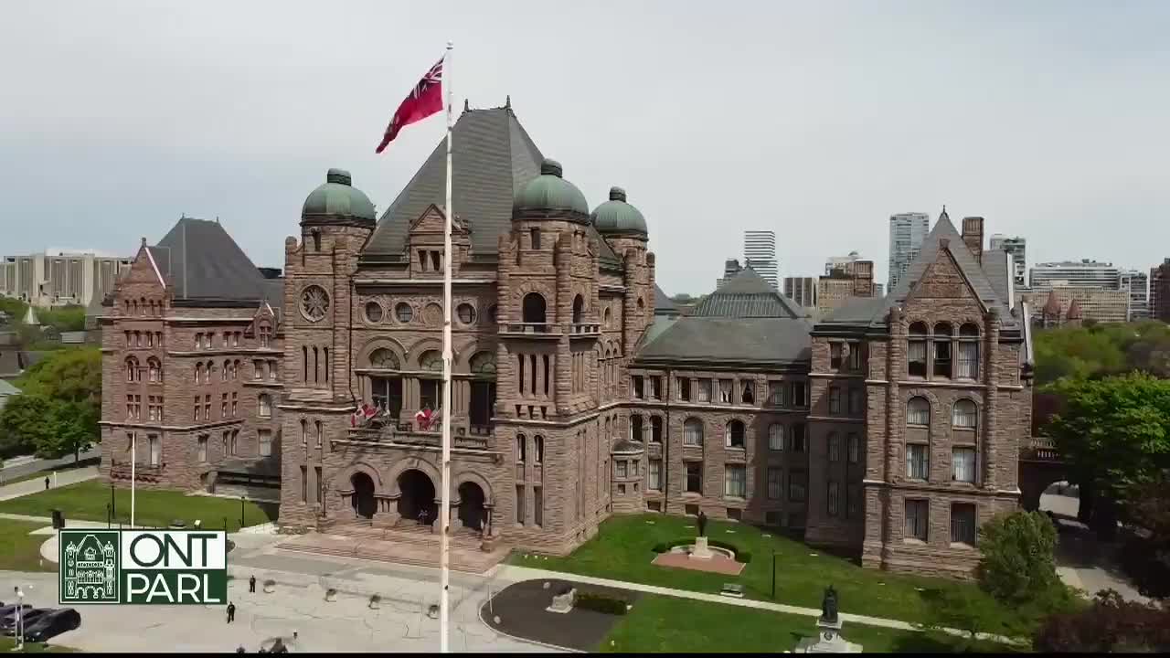 The red and white flag atop the Ontario Legislative Assembly flutters gently in the breeze. The stone facade of the building stands imposing against a backdrop of city skyscrapers.
