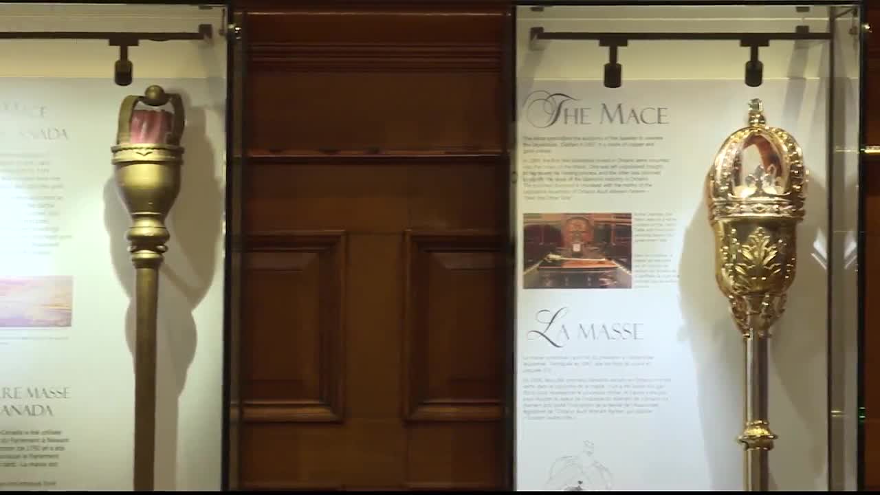 Two ornate maces, likely from the Legislative Assembly of Ontario, are displayed in glass cases. They stand tall, flanking a dark wooden door.
