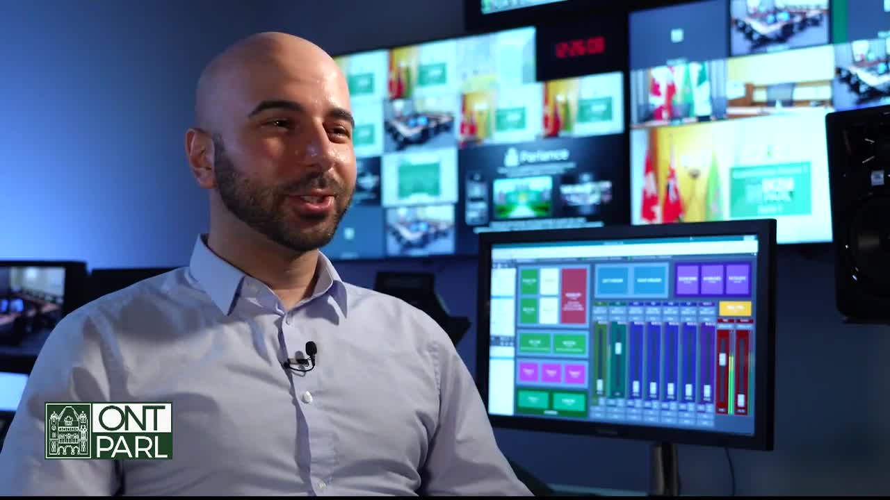 A man with a microphone is speaking, presumably for the Legislative Assembly of Ontario. Behind him, monitors display feeds and the time, along with the Parliament's logo.
