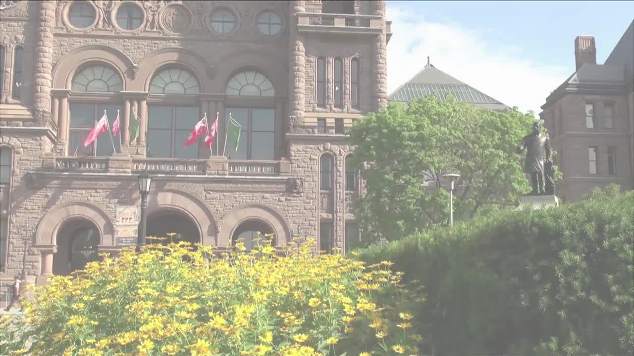 The Ontario Legislative Assembly building stands tall, with flags waving in the breeze. Yellow flowers bloom in the foreground, adding a splash of color to the scene.
