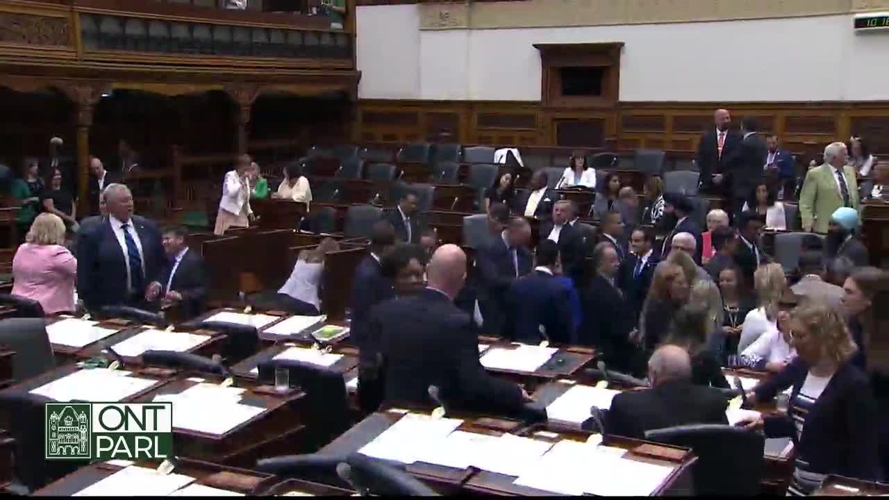 People in suits mill about the Legislative Assembly of Ontario chamber, some chatting, others heading toward their seats. A few individuals are standing near the desks, engaged in what looks like a lively discussion.

