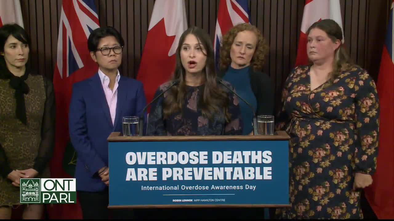 A woman at the podium speaks into a microphone, her mouth open as she addresses the room, while others stand behind her, watching. The Legislative Assembly of Ontario is in the background, with flags of Canada and Ontario visible.
