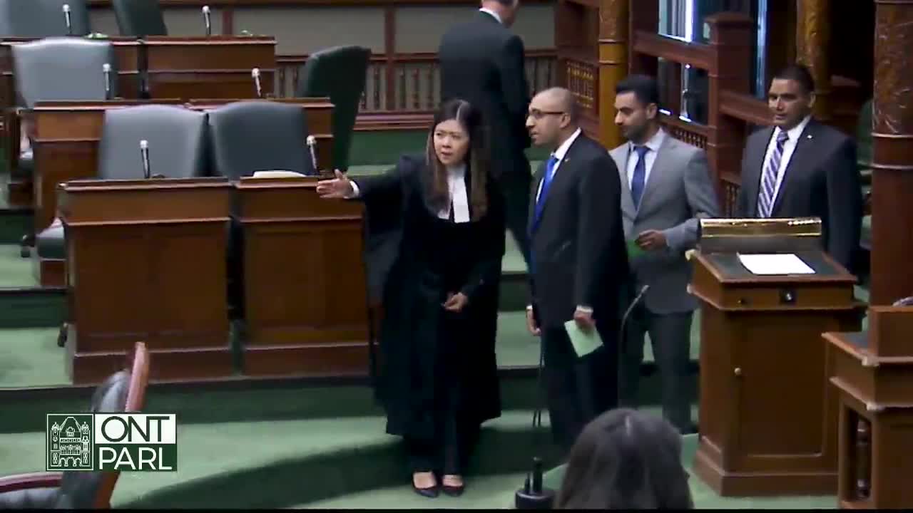 A woman in a black robe gestures with her right arm, addressing a group of men in suits. They stand near a podium in what appears to be the Legislative Assembly of Ontario.
