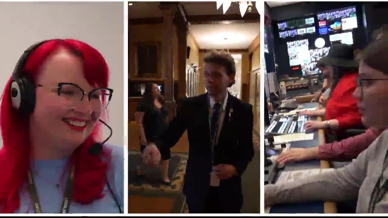 A woman with bright red hair and a headset smiles, likely broadcasting from the Legislative Assembly of Ontario. In the hallway, a man in a suit gestures as he walks, while behind him, a woman follows.
