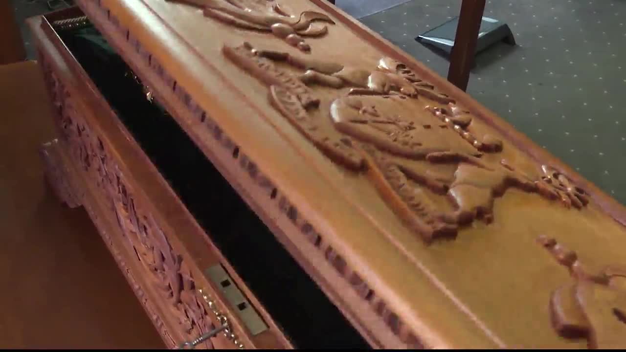 The lid of the wooden casket, ornately carved with figures, is slightly ajar, revealing the dark interior. A metal clasp is visible, and the casket appears to be in a room with patterned carpeting.

