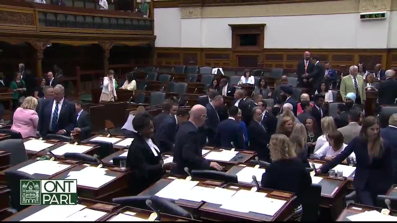 People in suits mill about the desks in the Legislative Assembly of Ontario, some chatting, others gathering papers. A few individuals are heading toward the back of the room.
