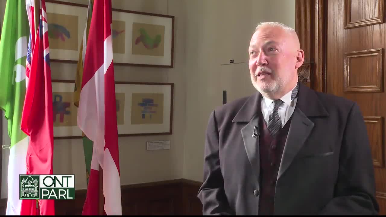 A man in a dark suit, likely at the Legislative Assembly of Ontario, is speaking, a microphone clipped to his lapel. Behind him, the Canadian flag and other flags hang, and the door is a rich, dark wood.
