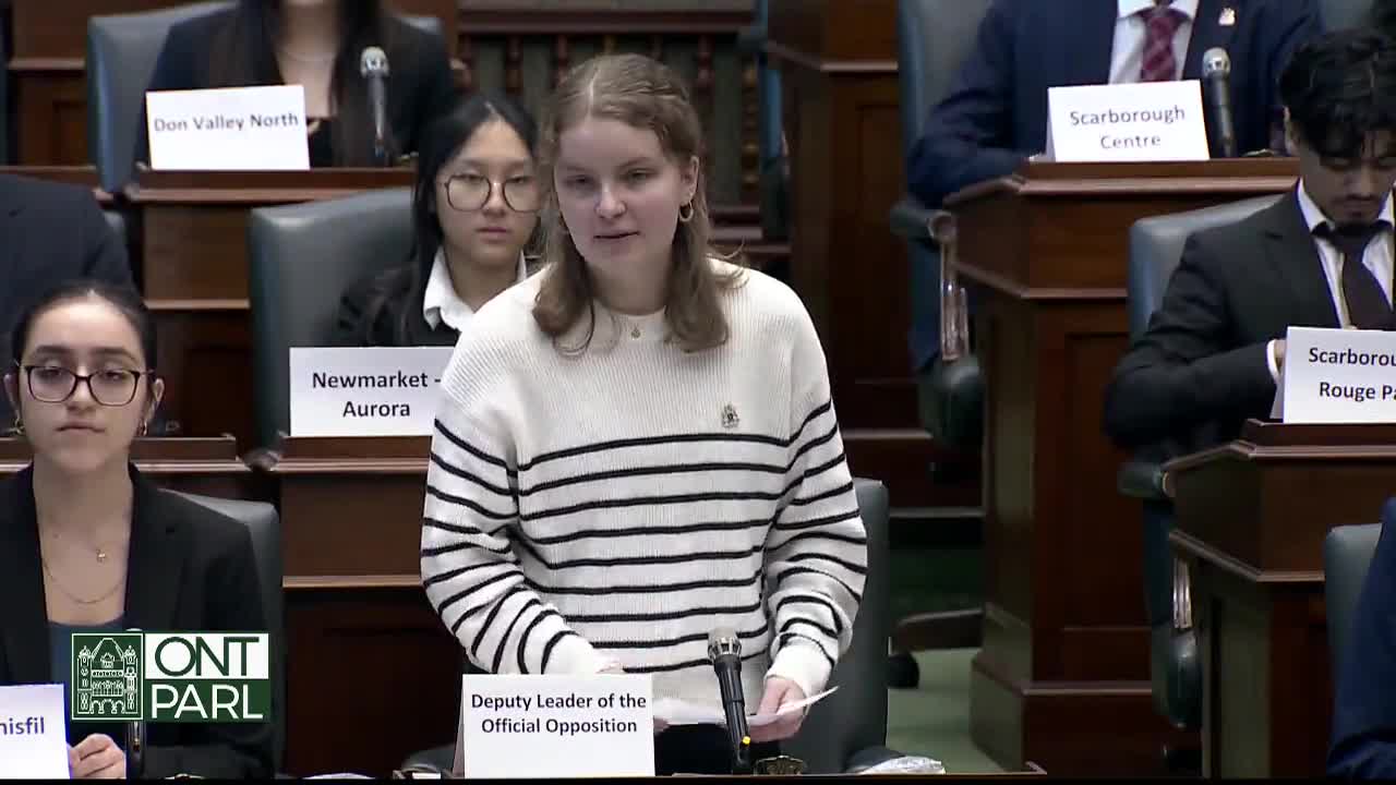 A young woman in a striped sweater stands at a podium, speaking into a microphone in the Legislative Assembly of Ontario. She holds a paper in her hand, while other members of the assembly sit behind her.
