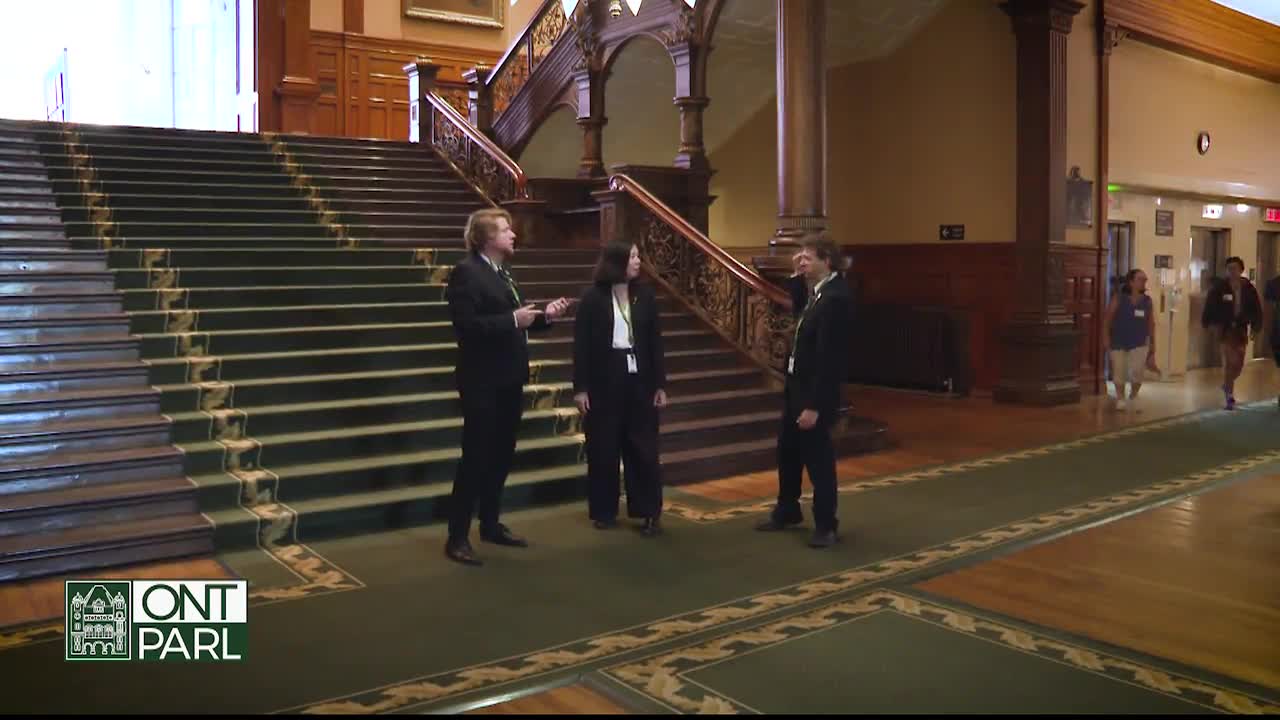 Three people in suits stand on a patterned carpet in the Ontario Legislative Assembly, near a grand staircase. The man on the left gestures as he speaks, while the other two listen.
