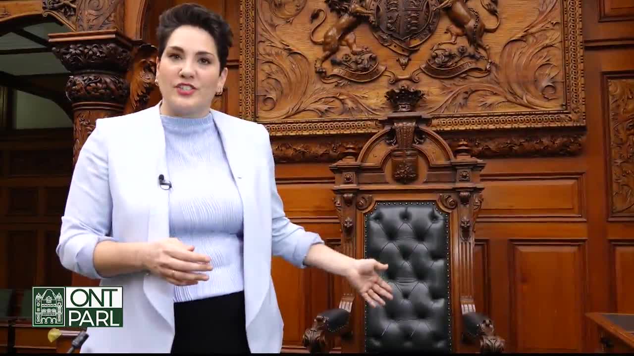 A woman in a light blue blazer gestures towards a large, ornate chair in the Legislative Assembly of Ontario. She's speaking, standing in front of the intricately carved wood paneling.
