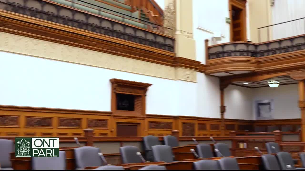 The interior of the Legislative Assembly of Ontario is visible, with rows of empty chairs facing the front.  Ornate wooden paneling and architectural details adorn the walls of the chamber.
