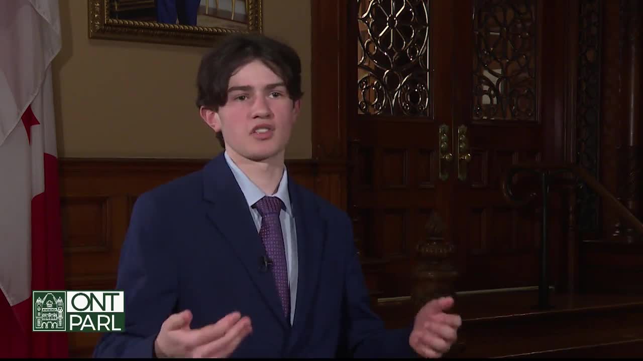 A young man in a suit gestures with his hands as he speaks, likely at the Legislative Assembly of Ontario. The Canadian flag is visible to his left, and ornate wooden doors are behind him.
