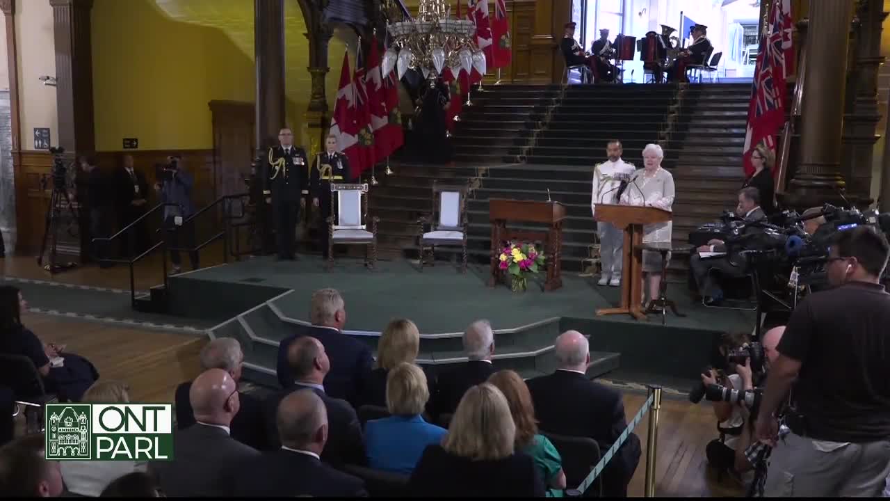 The Lieutenant Governor of Ontario stands at a podium, speaking in the Legislative Assembly. Canadian flags and a small band are positioned behind her on the grand staircase.
