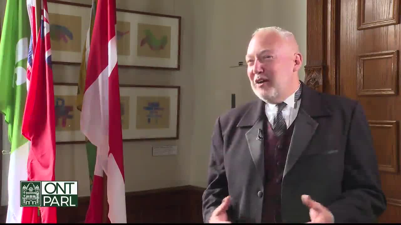 A man in a dark suit speaks animatedly, a small microphone clipped to his lapel. Behind him, flags of Ontario and Canada stand still in the hallway of the Legislative Assembly.
