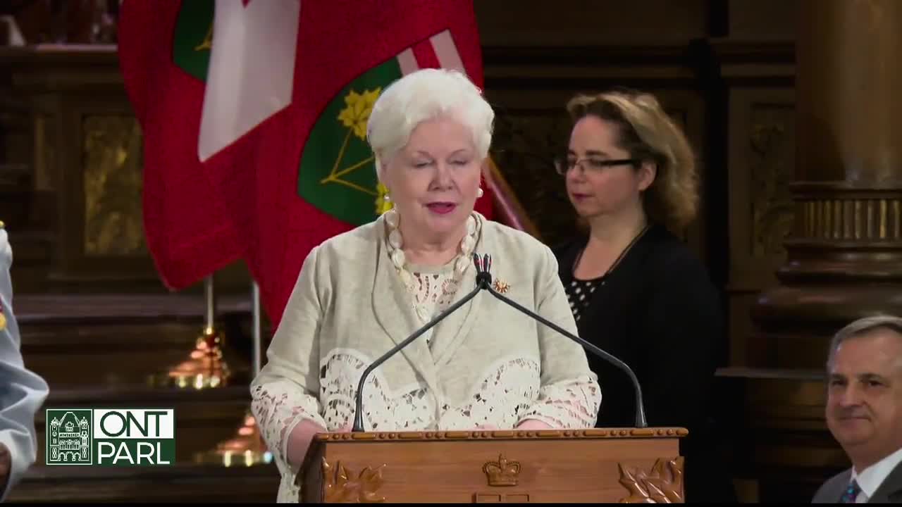 A woman in a cream-colored jacket speaks from behind a wooden podium, the Ontario flag visible behind her. Another woman stands to her side, listening.
