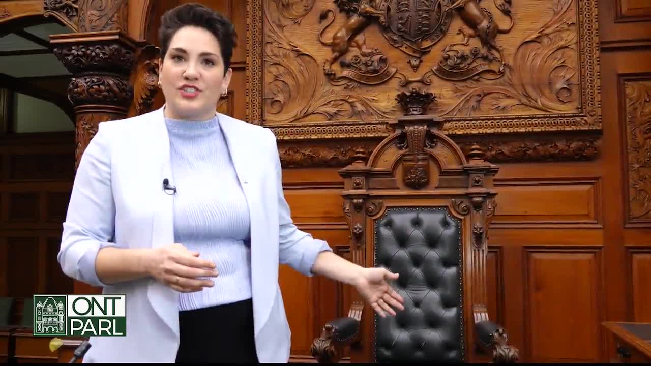 A woman in a light blue blazer gestures towards a large, ornate chair in the Legislative Assembly of Ontario. The rich wood paneling and carved crest behind her create a formal setting.
