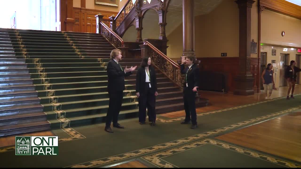A man in a suit gestures emphatically as he speaks to two people in the Legislative Assembly of Ontario. Further down the hall, people walk toward the elevators.
