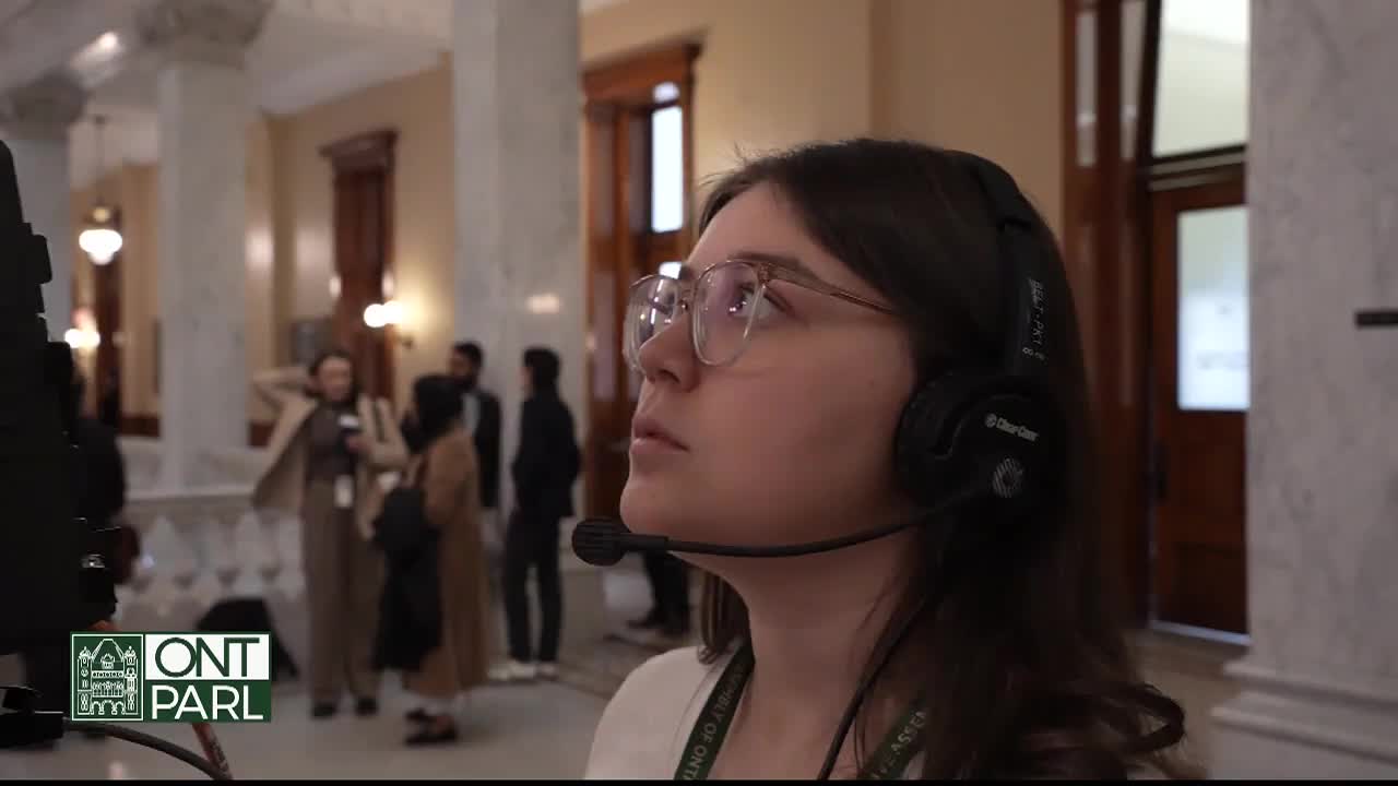 A woman wearing glasses and a headset gazes upward, likely focused on something out of frame. The scene unfolds within the grand architecture of the Legislative Assembly of Ontario, where others stand and converse in the background.
