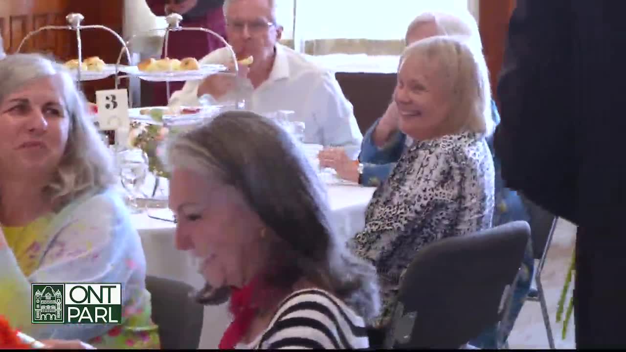 At a table in the Legislative Assembly of Ontario, people are smiling and chatting. A woman in a black and white striped shirt laughs, while others enjoy what looks like a tea service.
