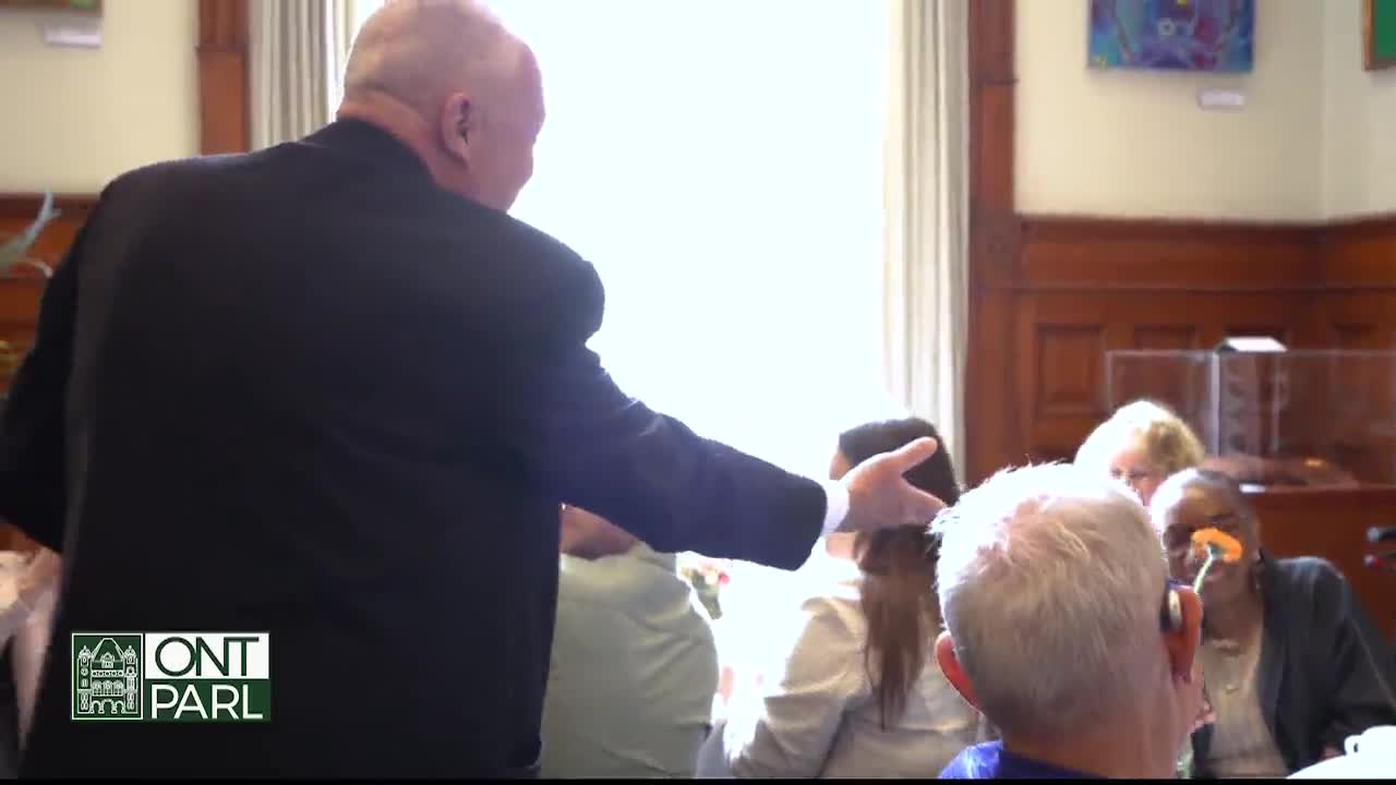 A man in a dark suit gestures toward a group of people seated in what looks like the Legislative Assembly of Ontario. The back of a man with white hair is in the foreground, and he's looking toward the speaker.
