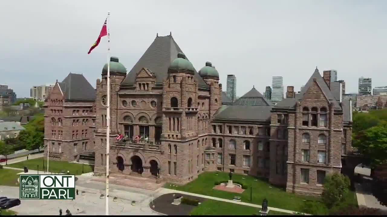 The Canadian flag and the Ontario flag are waving from the top of the Legislative Assembly building. The stone facade of the parliament building stands out against the backdrop of the Toronto skyline.
