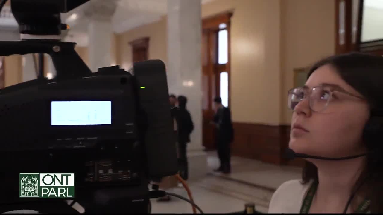 The camera operator, wearing headphones, stares intently at something off-screen, likely capturing the scene at the Legislative Assembly of Ontario. A few figures in dark suits stand in the background, near a large window.
