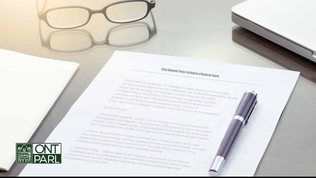 A document, likely a party statement from the Legislative Assembly of Ontario, rests on a desk, with a pen lying across it. Above, a pair of glasses sits, reflecting the light.
