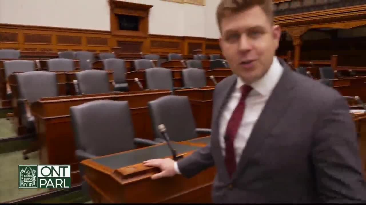 A man in a suit stands in the Legislative Assembly of Ontario, gesturing towards the empty seats. The camera focuses on him as he speaks, the wood paneling and rows of chairs providing the backdrop.
