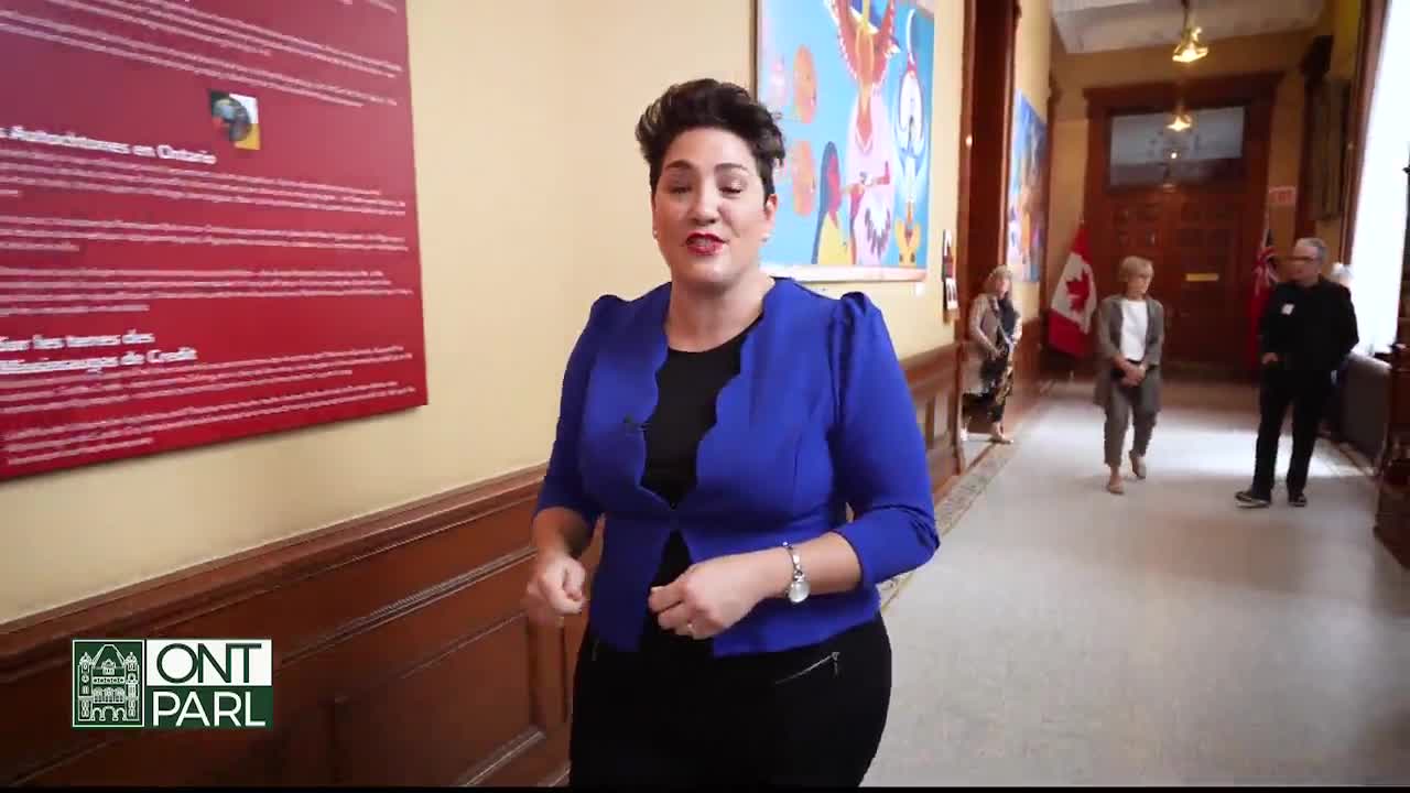 A woman in a blue jacket speaks directly to the camera in a hallway of the Legislative Assembly of Ontario. Behind her, a few people walk towards the camera, and a Canadian flag hangs on the wall.
