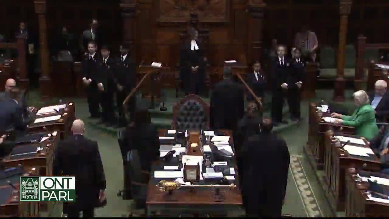 Inside the Legislative Assembly of Ontario, people in dark suits are walking around the chamber. A group of men in uniform stand at the back, while others are seated at their desks.
Inside the Legislative Assembly of Ontario, people in dark suits are walking around the chamber. A group of men in uniform stand at the back, while others are seated at their desks.
