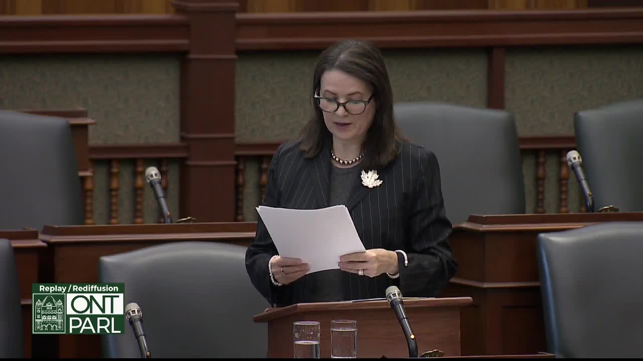 A woman in a pinstripe suit stands at a podium, reading from a document in the Legislative Assembly of Ontario. Her lips move as she speaks, and the paper in her hands is slightly rustling.
